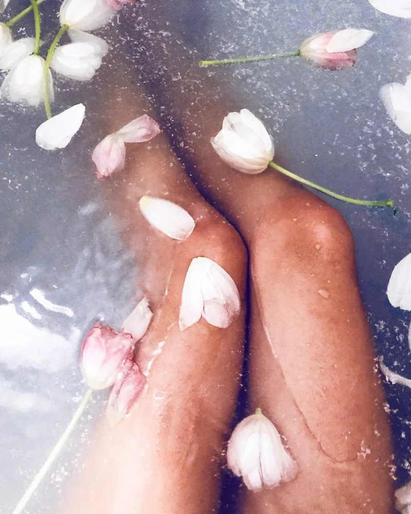 A person relaxing in a bath with flower petals, showcasing smooth legs after a Brazilian wax treatment.